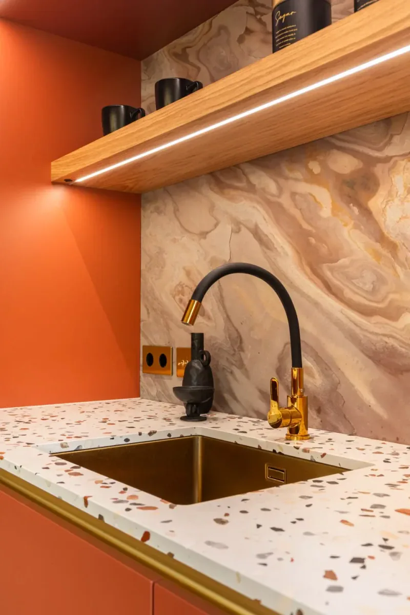 Photo detail of a brass sink and tap on a terrazzo worktop with a marble splashback and lit oak shelf